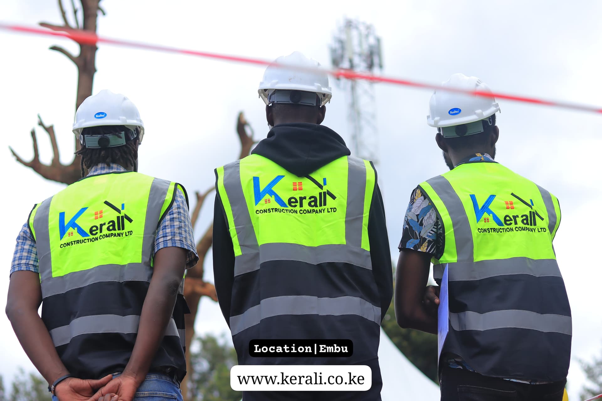 Three Kerali Construction workers photographed from behind at a site in Embu, in hi-vis vests printed with the company logo and white hard hats.